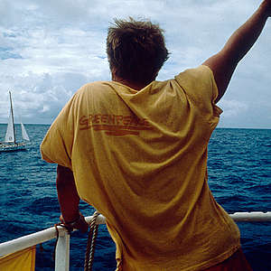 Crewmember MV Greenpeace waving to Vega during Pacific Peace Tour. 

Greenpeace sailing boat 'Vega' and ship "MV Greenpeace" during campaign against nuclear testing in the Pacific.
Previous to French nuclear test site protest in the Pacific, the MV Greenpeace and flotilla return to New Zealand to replace Rainbow Warrior I that was bombed in Auckland. The refit of the ship prior to departure was done at Oranjewerf, which is in Nieuwendam, to the NE of Amsterdam. The Inmarsat dome was installed in the Antilles and some engine maintenance, following the Atlantic crossing.