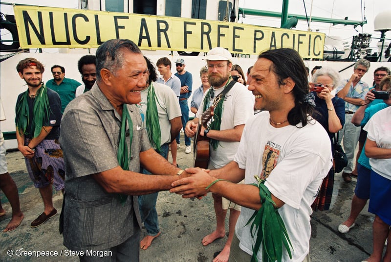 Deputy Prime Minister of the Cook Islands, Inatio Akaruru, greets SV Rainbow Warrior II crew member Jean-Marc Van Chinh (France) with Greenpeace Nuclear Campaigner Steve Sawyer (USA) playing guitar, and crew member Philip Pupuka (Solomon Islands) to the left of Inatio Akaruru