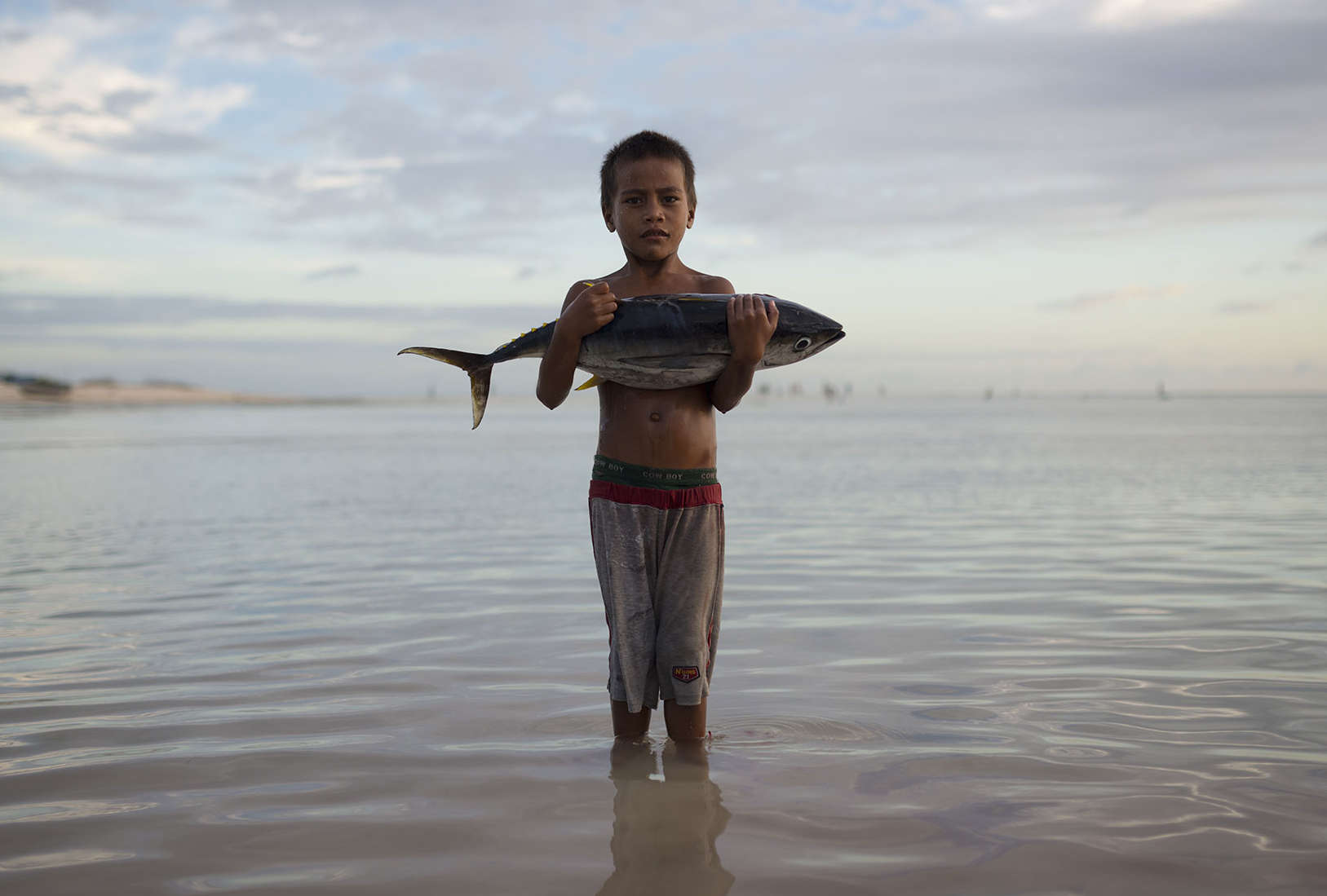 A young boy stands knee-deep in the ocean at dusk, looking directly into the camera and holding in his arms a large tuna fish.
