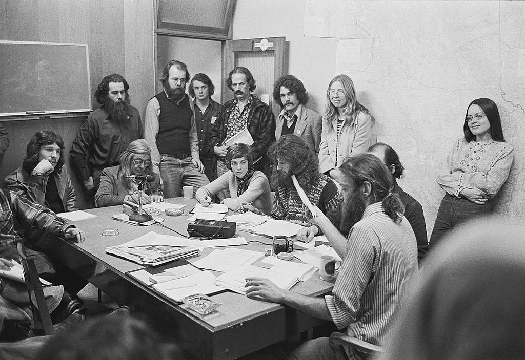 The first public Greenpeace office at 2007 West 4th Avenue in Vancouver, BC, Canada. Bob Hunter sits at the head of the table, Bree Drummond leans against the wall, Rod Marining, with long hair and beard sits to Hunter's right, Alan Clapp stands in the doorway with a file folder, and Henry Payne sits at the far end of the table with a headband. © Greenpeace / Rex Weyler