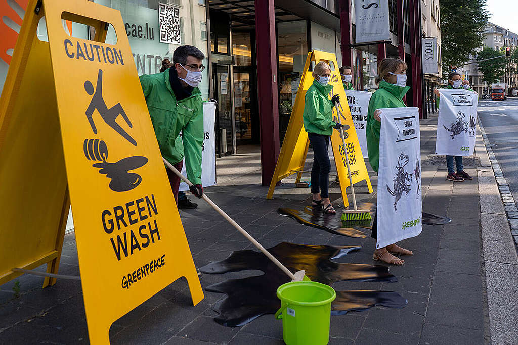 Greenpeace Luxembourg activists use human size yellow slipper floor warning signs and mops to protest continued fossil fuel investment in Luxembourg.