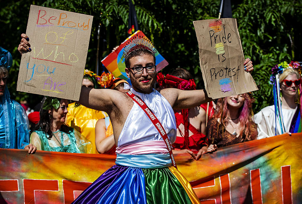 A person holds signs that read 'Be Proud of Who You Are' and 'Pride is Protest'. © Marten van Dijl / Greenpeace Picture showing a Queers4Climate activists standing against climate breakdown and holding two signs reading "Be proud of who you are" and "Pride is Protest".