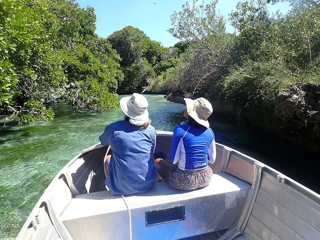 Two women with blue shirts and white hats sit on the end of a white boat floating down a body of water. © Emma Mederic Two women with blue shirts and white hats sit on the end of a white boat floating down a body of water. © Emma Mederic