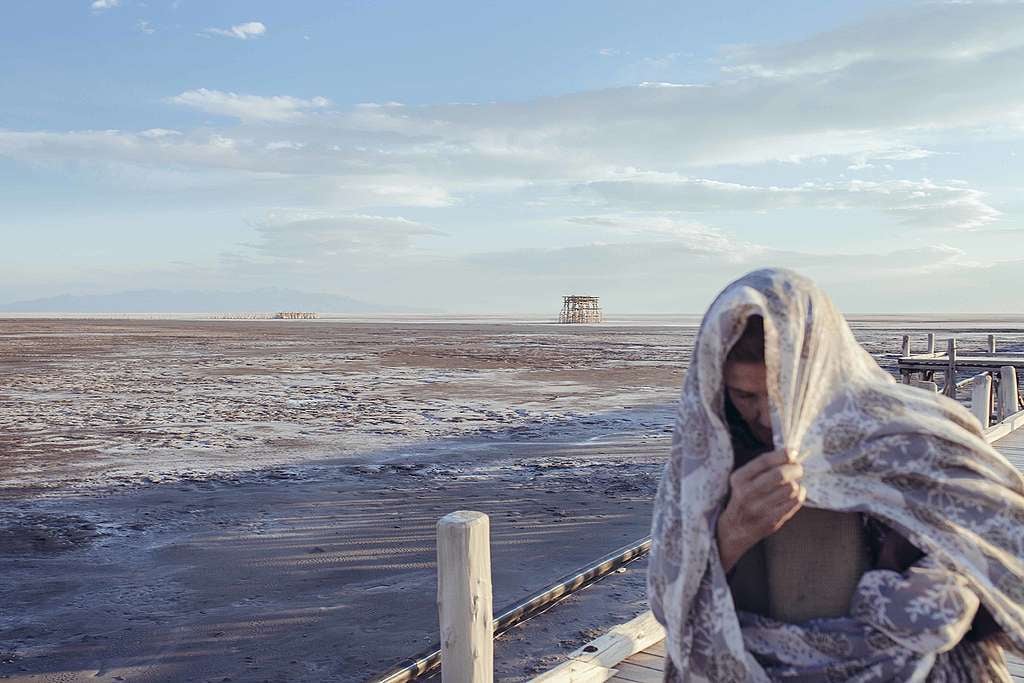 An elderly woman covering her head in a scarf is walking past the coast of a port town in Iran. The lake is very dry and no water is visible.