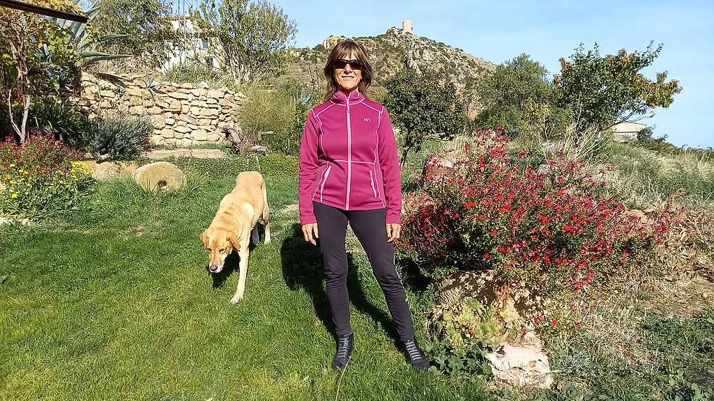 Pilar Torres L’era (Santa Eulalia la Mayor, Loporzano, Huesca, Aragón) stands in a garden looking to camera as her dog walks by.