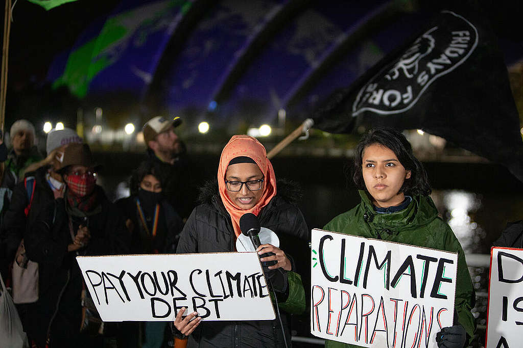 A girl with a head scarf speaking into a mic while holding a placard that says Pay Your Cimate Debt. Another girl is standing next to her holding a placard that says Climate Reparations. A girl with a head scarf speaking into a mic while holding a placard that says Pay Your Cimate Debt. Another girl is standing next to her holding a placard that says Climate Reparations.