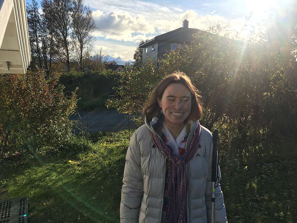 A white person with shoulder length hair stands in winter greenery outdoors, smiling. They're holding their white cane, and mountains are visible behind them.