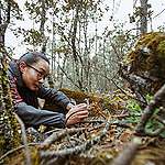 A girl with long hair and glasses laying on the ground with a camera in her hand, in a forest