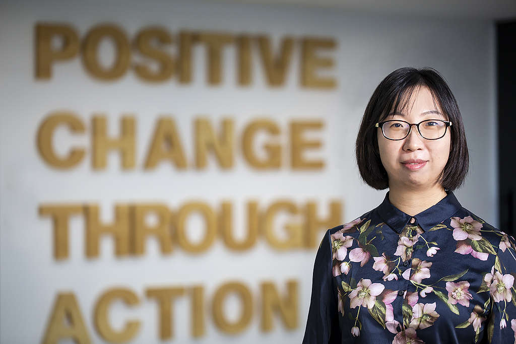 A girl with shoulder length hair and glasses looking at the camera. Behind her are the words Positive Change Through Action on the wall.