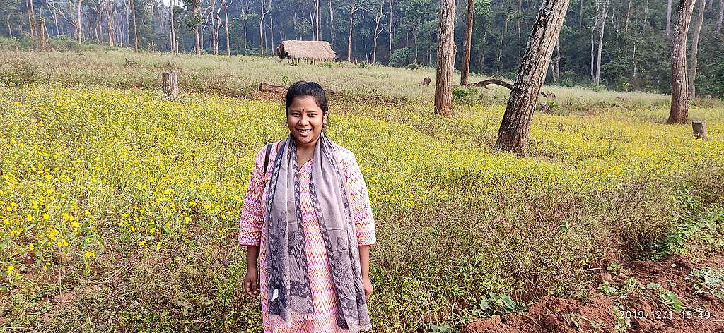 A girl standing in a field of grass, smiling. There is a small hut and trees in the background