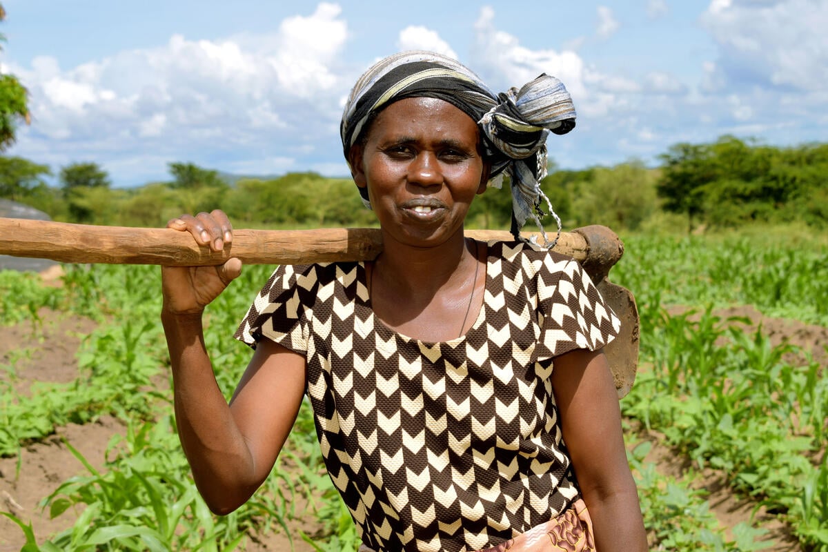 Smiling black woman in graphic brown and white patterned dress and stripy headscarf, ecological farmer Stella Muthama in Kenya, looks at the camera with her ploughing tool over her shoulder and crops in the background.