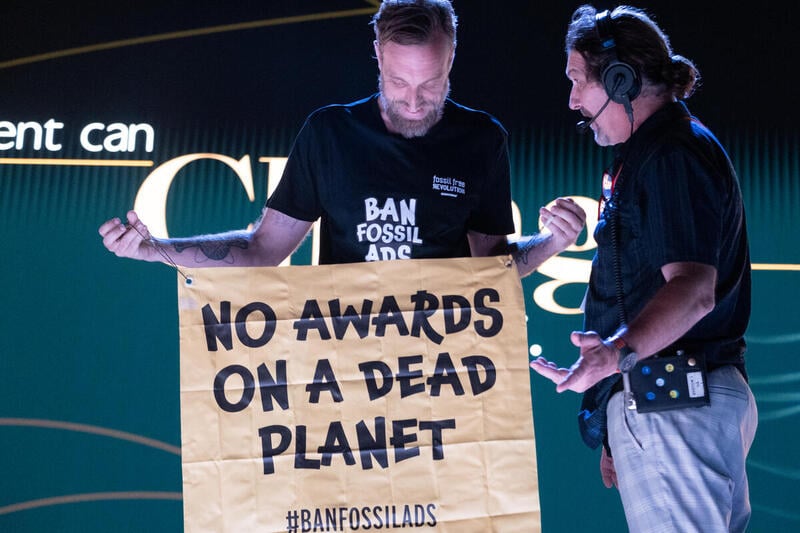 White man with brown beard wears black t-shirt that says Ban Fossil Ads in white writing. Holds a sign saying No Awards On A Dead Planet. Man to his left gestures as if asking, what's going on? A Greenpeace France activist and former winner and jury at the Cannes Lions awards - the famous advertising festival - interrupted the opening ceremony in Cannes to give back an award he won for an airline company advert.