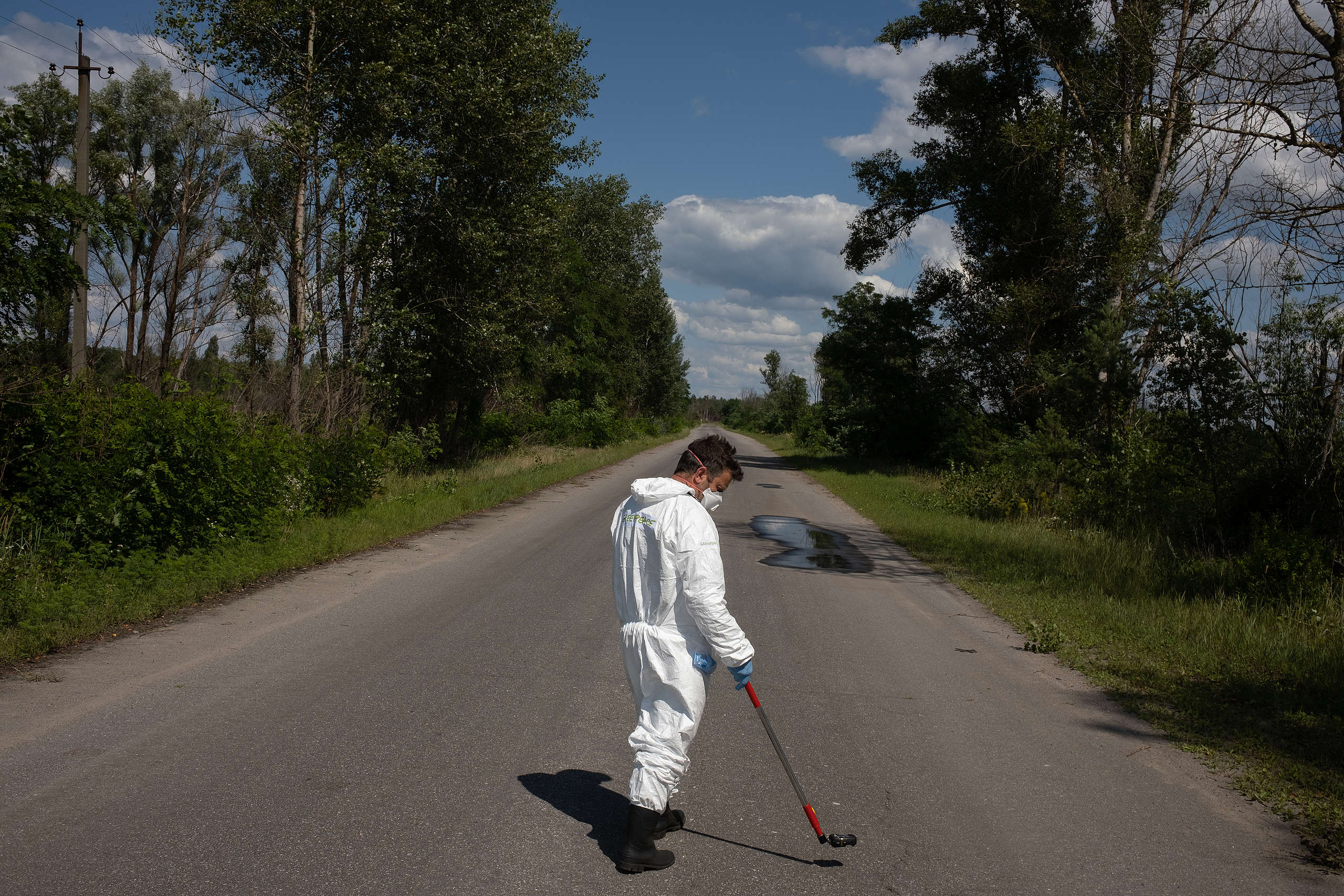 Person in a white protective overall and mask uses radiation detection equipment along a road lined with trees in the Chornobyl nuclear exclusion zone.