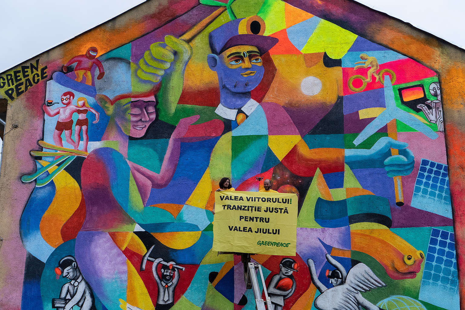 Lively colourful street art on the wall of a brick building in Romania pictures the transition from a community centred on mining to a one producing its own clean energy, with solar panels and people riding bikes instead of wielding pick axes. Yellow Greenpeace banner with black writing calls for a just transition for the Jui Valley region (Valea Jiului in Romanian).