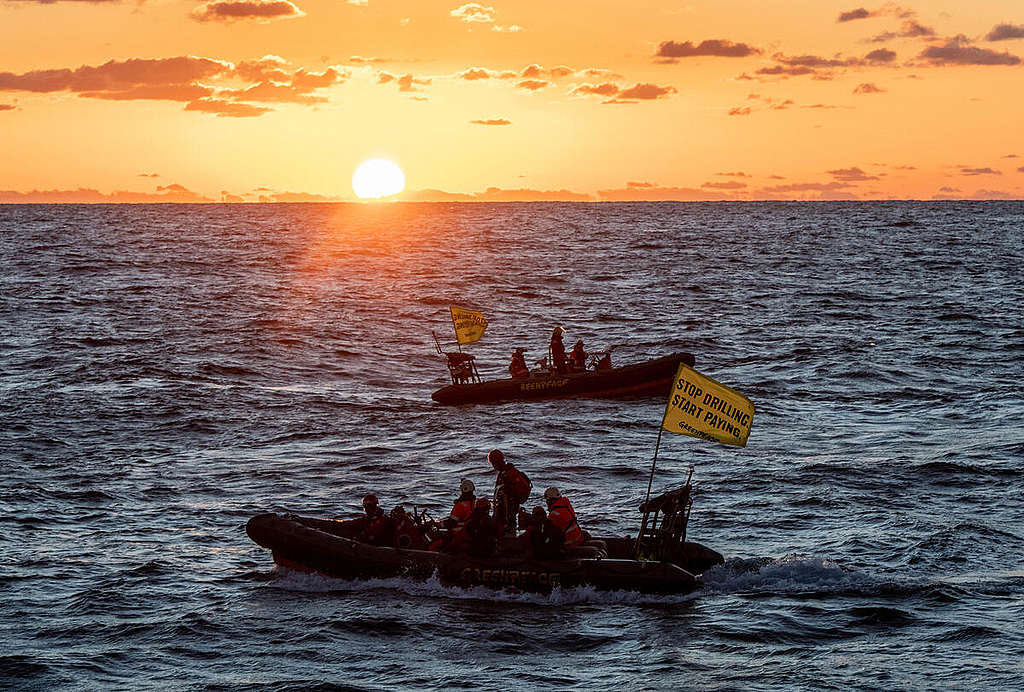 Sun on the horizon as Greenpeace activists from climate-hit countries on a small boat head out to floating Shell platform that is en route to major oilfield with message: ‘STOP DRILLING. START PAYING.’
