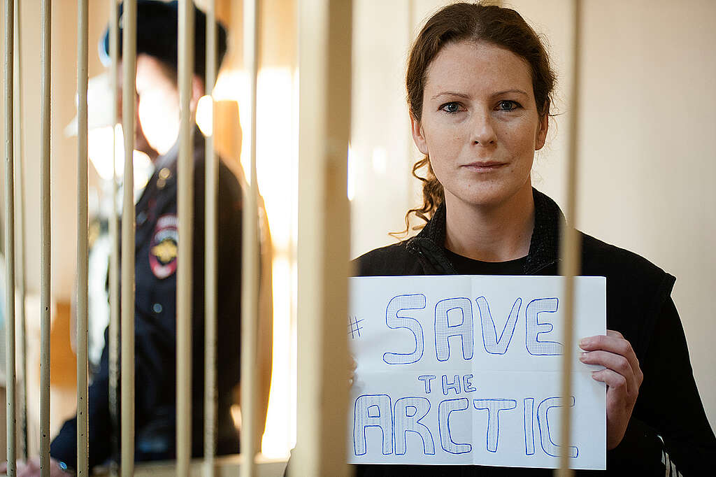 Ana Paula Alminhana Maciel Detention Hearing in St. Petersburg. © Vladimir Baryshev / Greenpeace At a detention Hearing in St. Petersburg, Russia. Activist Ana Paula from Brazil is pictured behind bars holding a handwritten sign saying #SaveTheArctic. A guard in uniform stands behind her © Vladimir Baryshev / Greenpeace