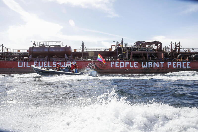 Small activist boat in front of a large red fossil fuel vessel with the words OIL FUELS WAR. PEOPLE WANT PEACE. painted in white