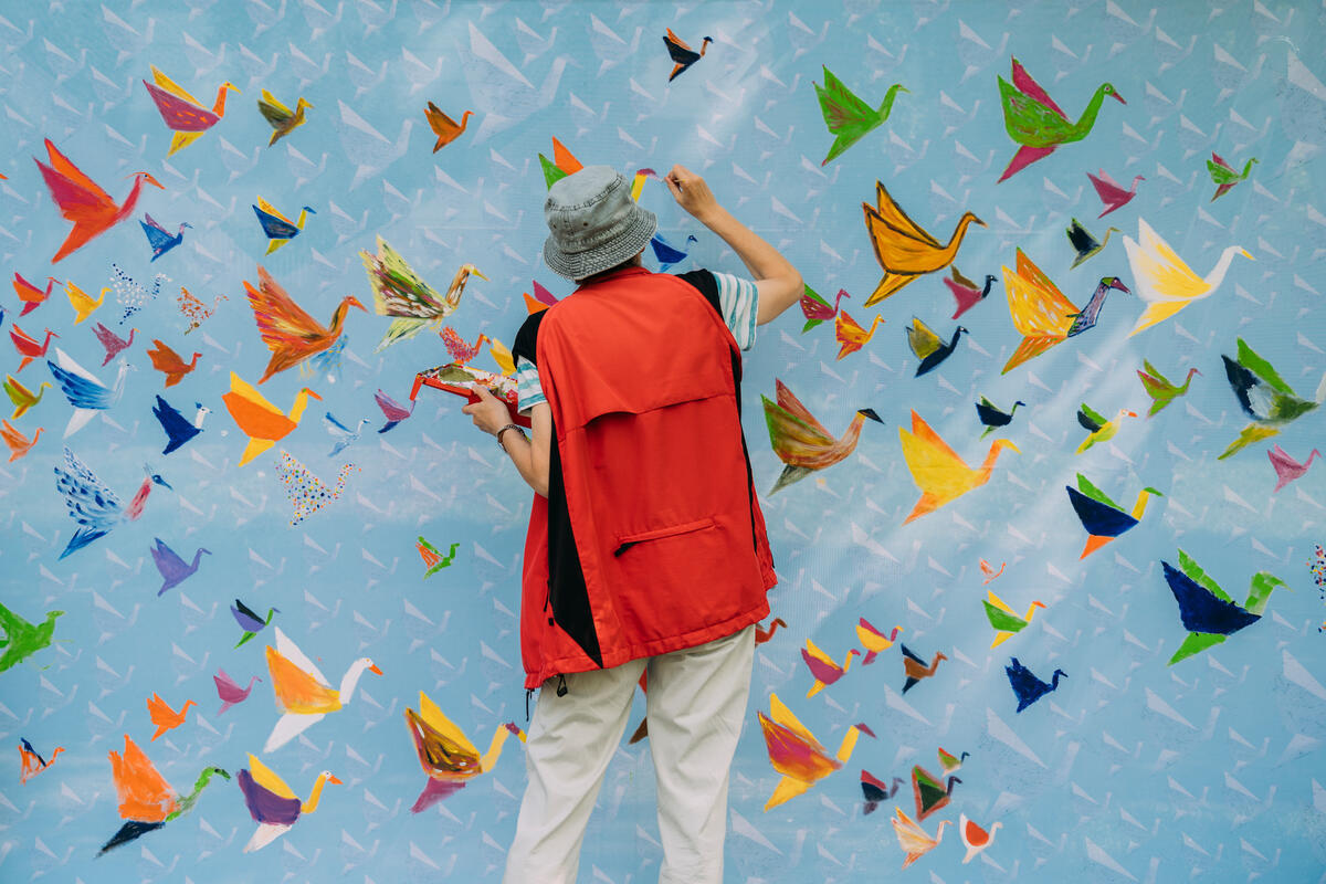 The Thousand Cranes Mural-Banner on Hiroshima Peace Memorial Day in Hamburg. © Jewgeni Roppel / Greenpeace Colourful birds on a blue background as artist in red jacket puts final touches to Thousand Cranes Mural-Banner on Hiroshima Peace Memorial Day in Hamburg. © Jewgeni Roppel / Greenpeace