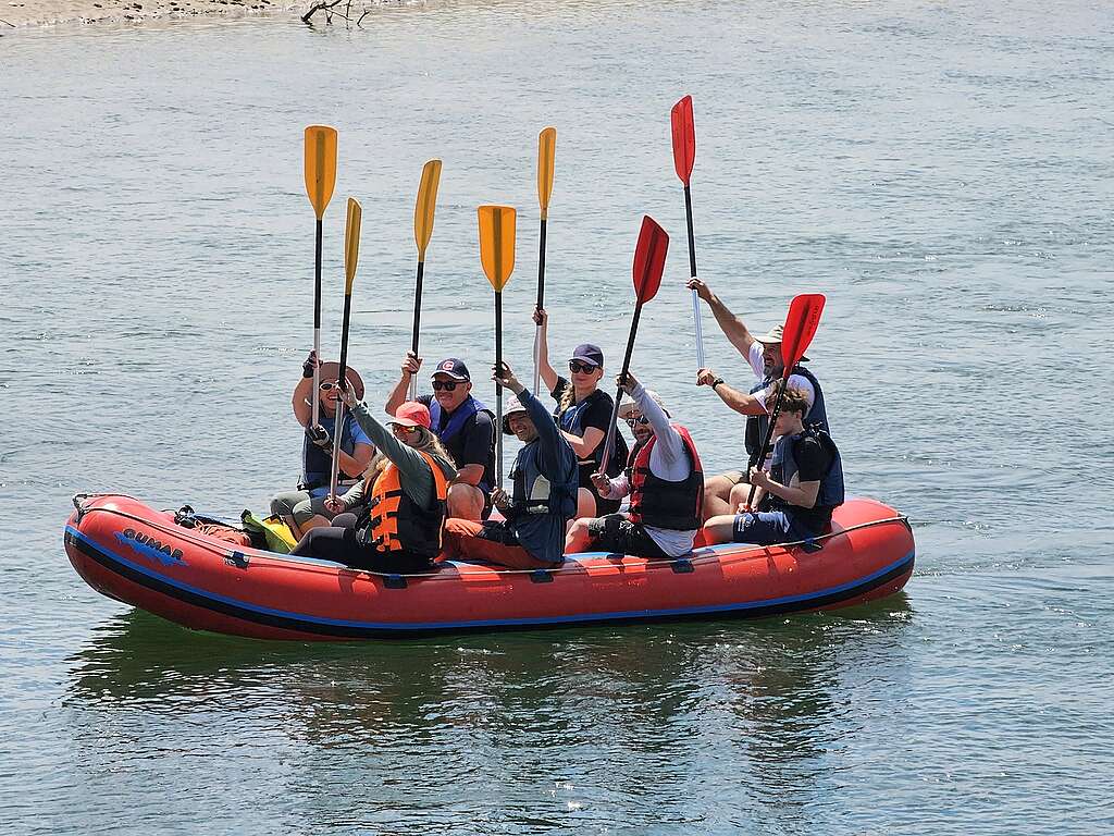 A group of people paddles a red inflatable raft along the Drava River, with colorful paddles raised above the boat.