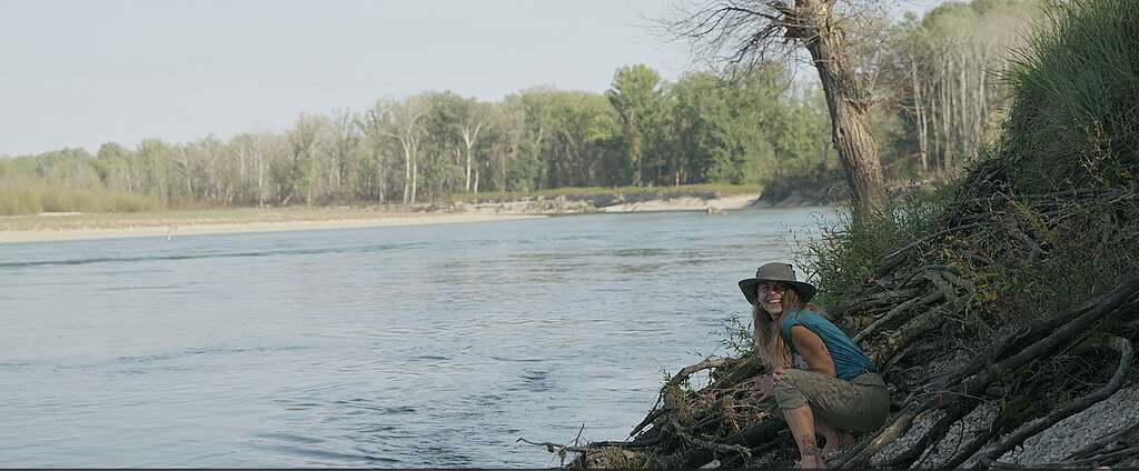 Kristina finding a small snake under the roots of a tree A smiling woman wearing a brimmed sunhat and teal top crouching near the exposed tree roots along the muddy bank of the Drava River. The wide river flows behind her under a pale sky with lush green trees lining the opposite shore.