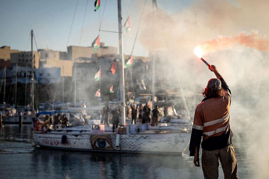 People board decorated sail boats at a port in Sicily, part of a peaceful flotilla sailing to break the siege of Gaza. Person in the foreground holds a bright flare above their head. © Max Cavallari / Greenpeace
