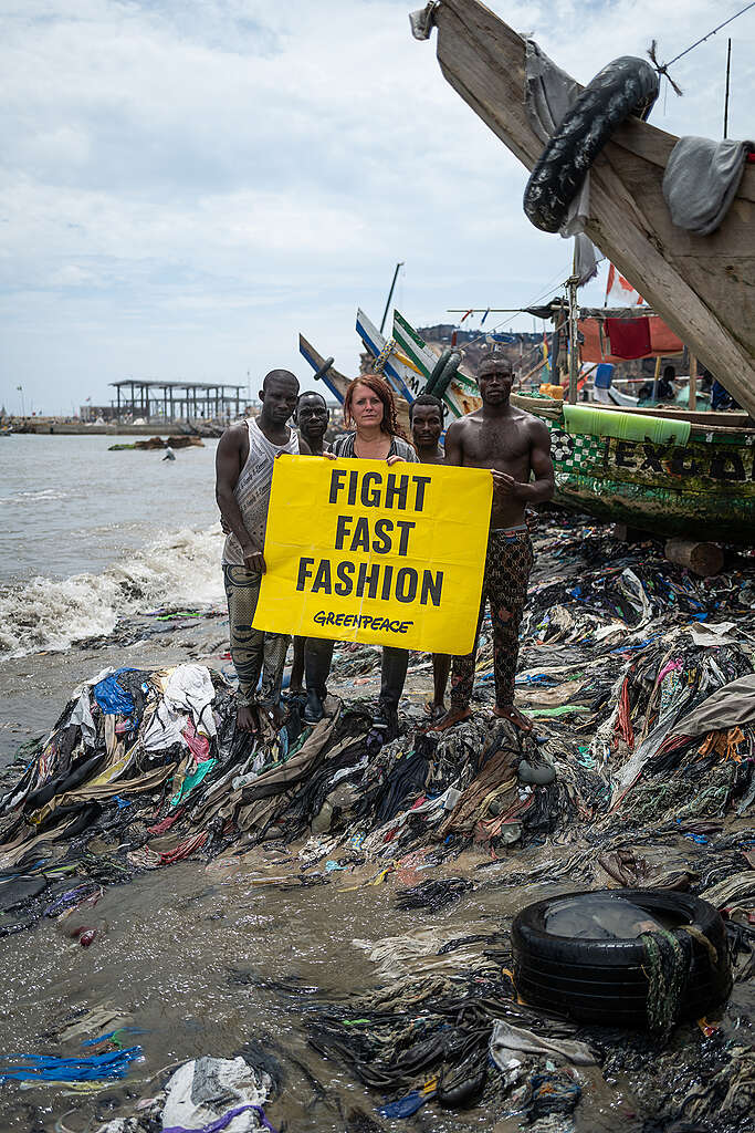 אישה וארבעה גברים מחזיקים שלט צהוב נגד תעשיית האופנה המהירה כשהם עומדים על פסולת טקסטיל בחוף הים. 