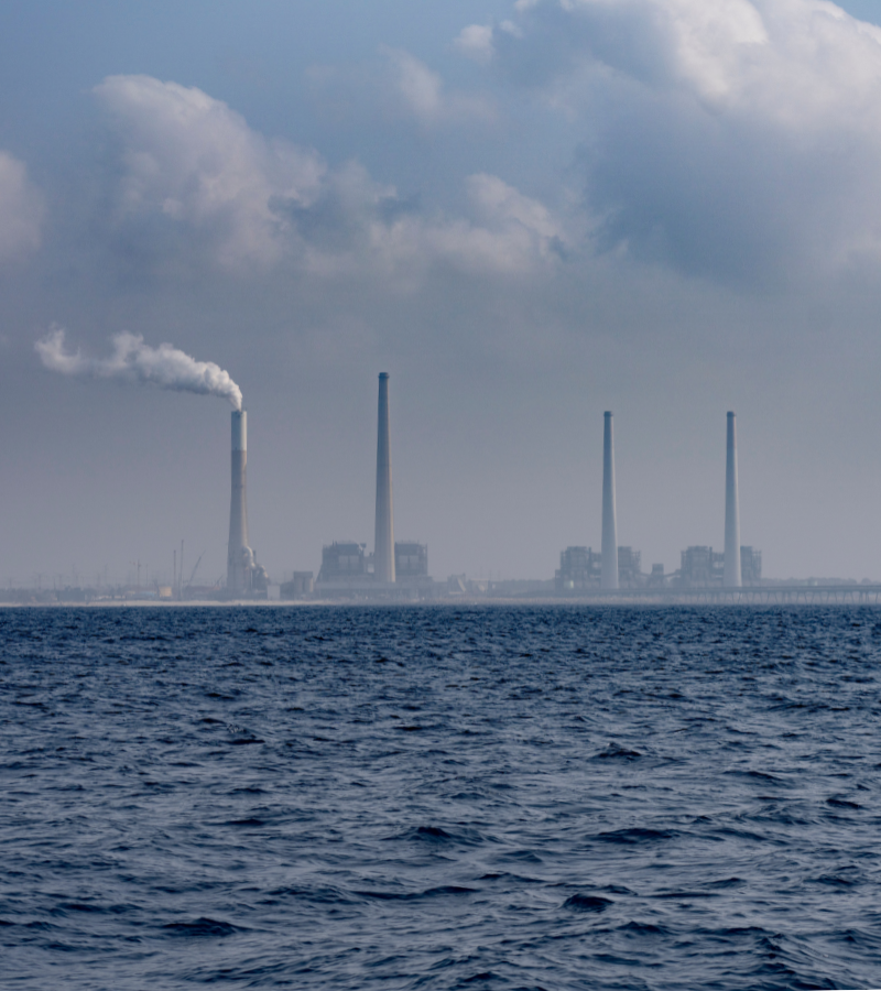 צילום של הים התיכון וברקע ארובות תחנת הכוח ״אורות רבין״ בחדרה עם עשן לבן שיוצא מאחת הארובות.