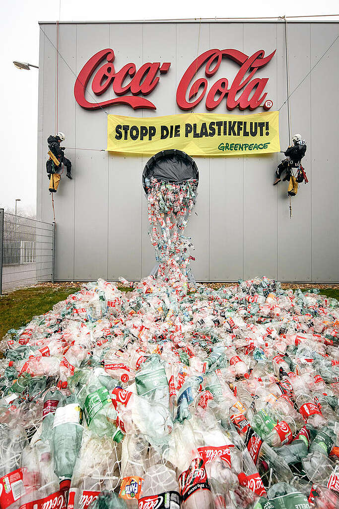 שני פעילי גרינפיס מטפסים על מפעל חברת קוקה-קולה ופורסים שלט צהוב עליו כתוב ״להפסיק את ייצור הפלסטיק״ כשמתוך המפעל יש מיצב של בקבוקי פלסטיק ריקים של קוקה-קולה.