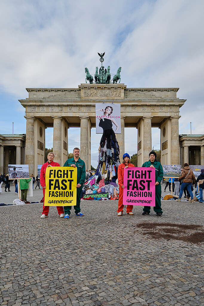 ארבעה פעילי גרינפיס עומדים על רקע מיצב נגד אופנה מהירה עם שלטים ״עצרו את האופנה המהירה״. 