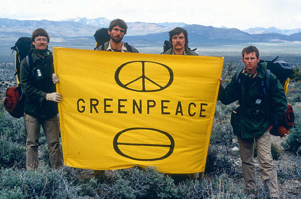 תמונה משנות השבעים של ארבעה גברים עם תיקי מטיילים מחזיקים שלט צהוב עליו כתוב גרינפיס וסמל השלום.