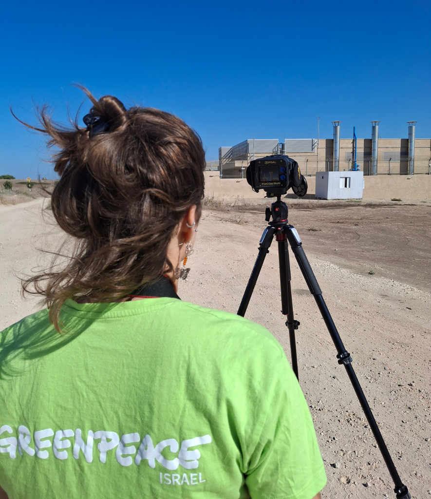 פעילת גרינפיס בחולצה ירוקה מצלמת עם מצלמה תרמית מפעל בישראל.