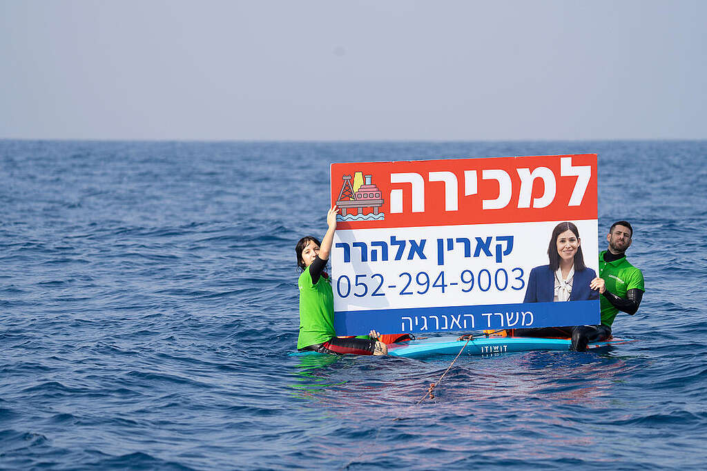שני פעילים של גרינפיס ישראל עם חולצות ירוקות עומדים בתוך הים ומחזיקים שלט עם תמונה של שרת האנרגיה דאז קארין אלהרר ועליו כתוב ״למכירה״.