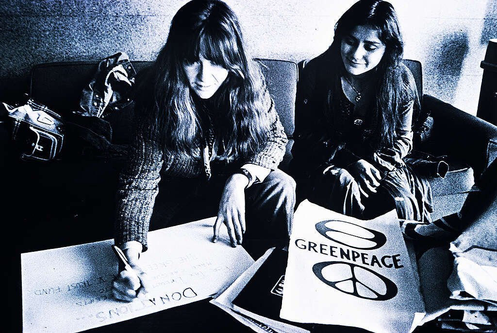 תמונה בשחור לבן של שתי נשים יושבות על ספה, אחת כותבת שלט בכתב יד ולצידו שלט שכתוב עליו גרינפיס עם סמל השלום