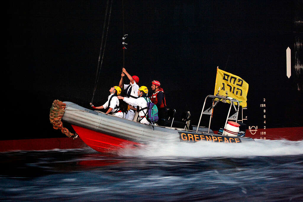 פעילי גרינפיס בתוך סירה מתנפחת עם דגל צהוב עליו כתוב ״פחם הורג״ עומדים לטפס על ספינה גדולה.
