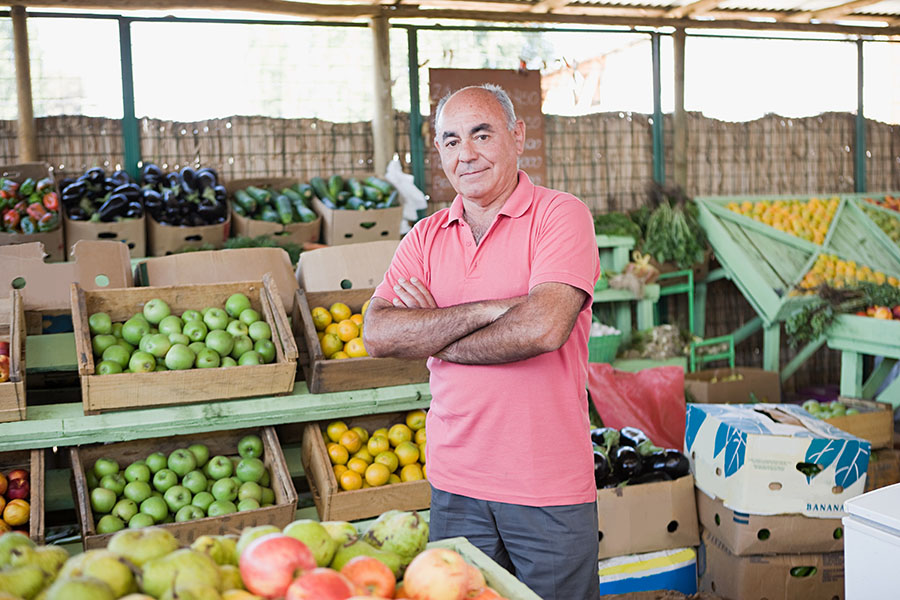 2c8ef152 Cheerful Man At A Farmer S Market With Produce 2026 09 22 04 26 23 Utc מוכר בשוק עומד בין דוכני פירות וירקות טריים
