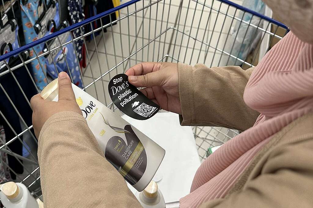 A woman with a Dove shampoo bottle in her shopping cart, featuring a "Stop Dove's Plastic Pollution" sticker that raises awareness.