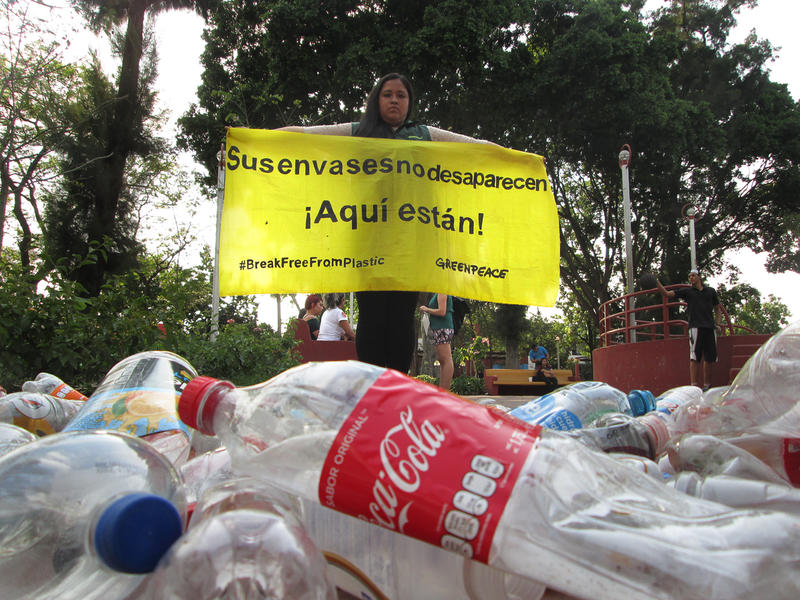  Botellas de plástico tiradas en el suelo y en el fondo una activista de Greenpeace con un banner que dice: Sus envases no desaparecen. ¡Aquí están!