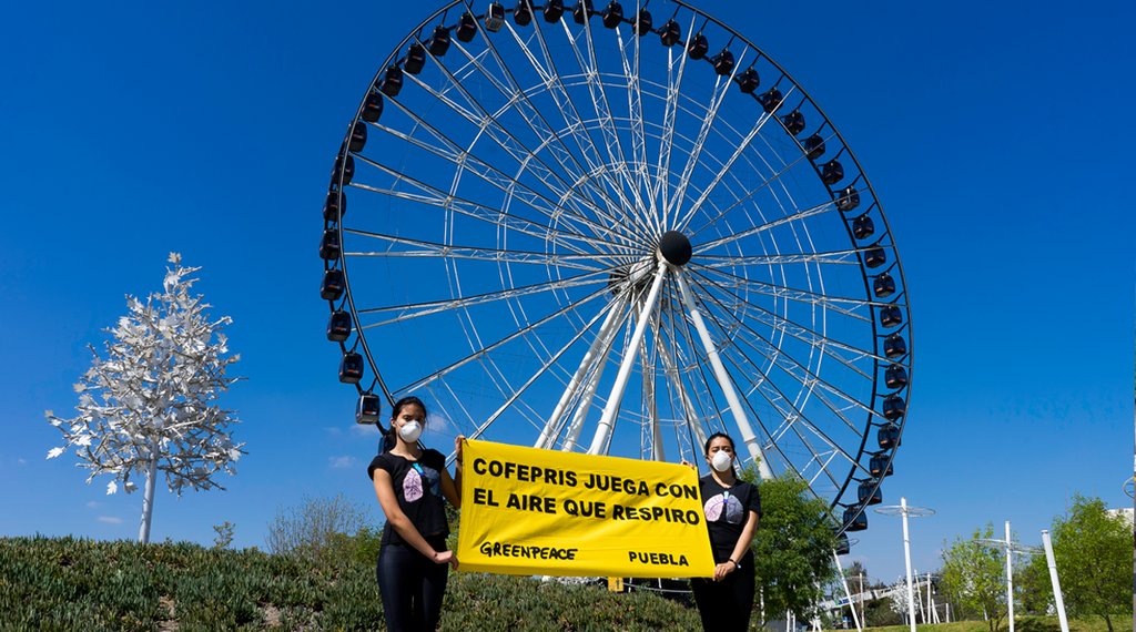 Dos voluntarias de Greenpeace México en Puebla usan cubrebocas y sostienen una manta con el mensaje “Cofepris juega con el aire que respiro”, como parte de una protesta por la mala calidad del aire.