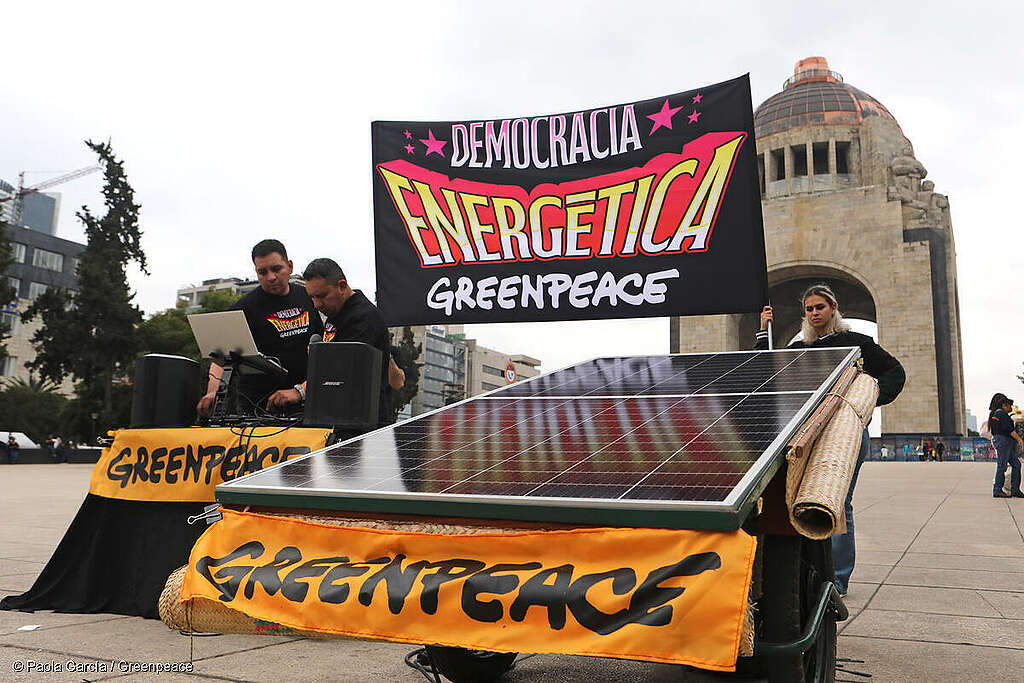 Activistas de Greenpeace México con un panel solar y una bandera que dice 'democracia energética' caminando frente al Monumento a la Revolución en CDMX, luchando por la justicia energética.