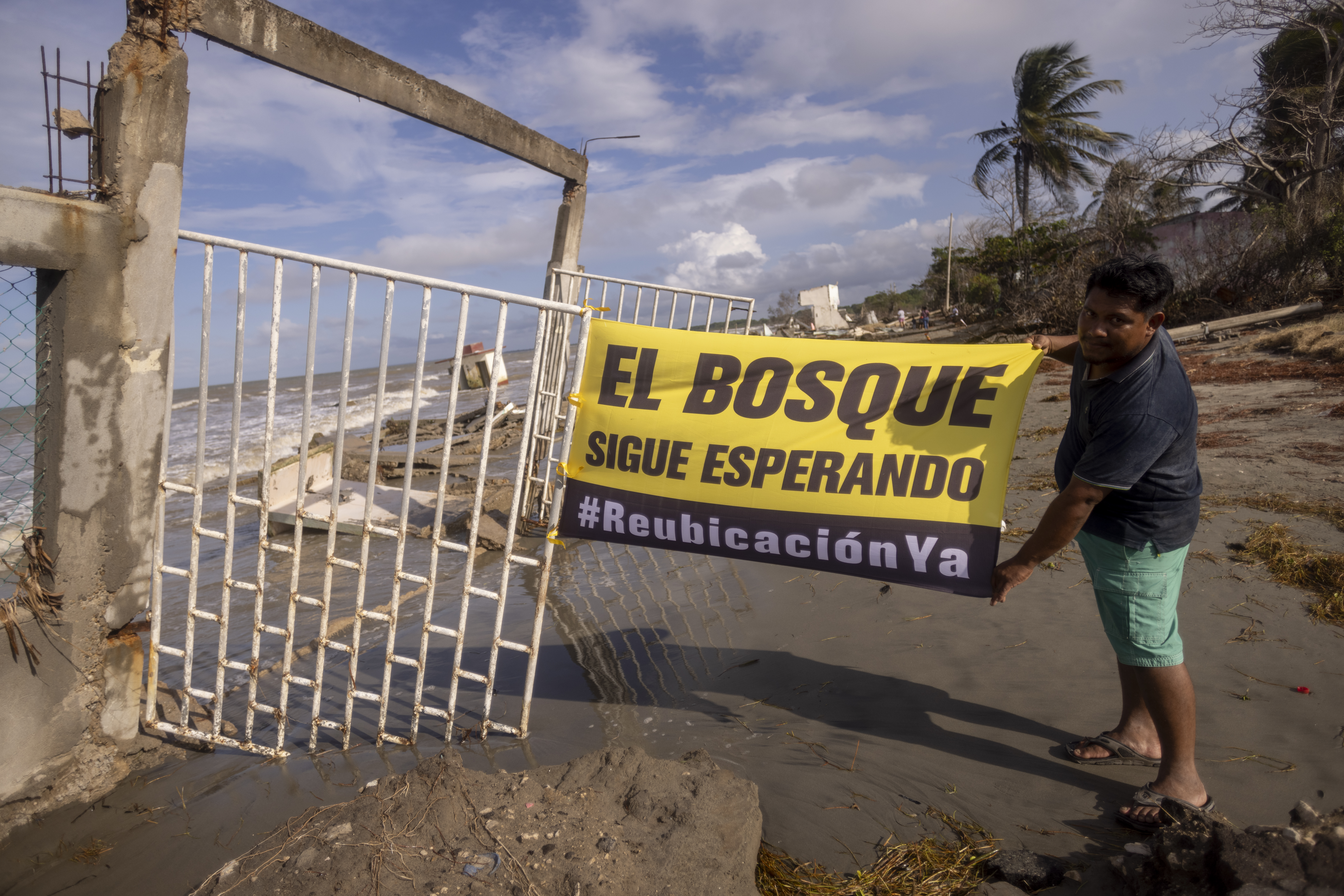 Habitante de la comunidad El Bosque sostiene una manta que dice “El bosque sigue esperando #ReubicaciónYa”, entre restos y daños por el desplazamiento climático.