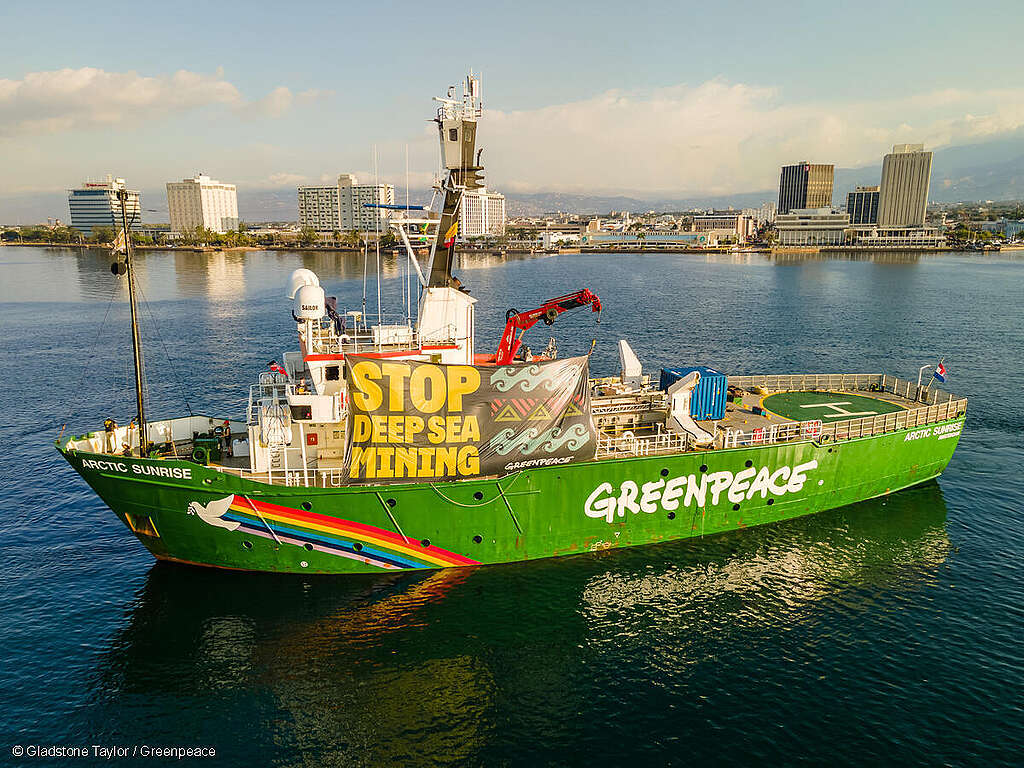 Fotografía del barco Arctic Sunrise iluminado por el sol, con colores vivos. En la cubierta cuelga una manta que dice “Alto a la minería en aguas profundas”.