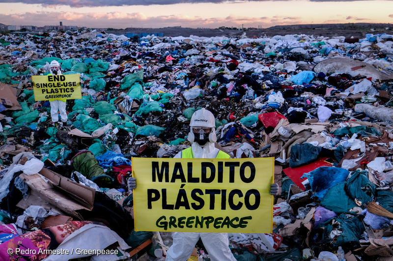 Dos activistas de Greenpeace con trajes y mascarillas de protección están de pie entre una montaña de plásticos y basura, sosteniendo mantas que dicen "Maldito plástico".
