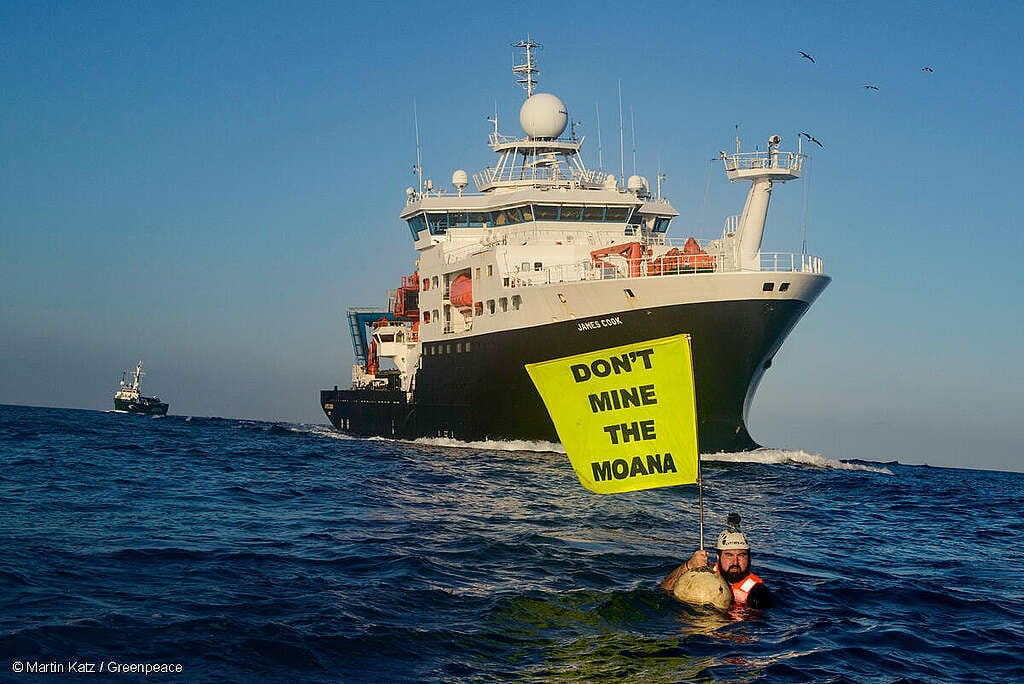 Activista de Greenpeace en medio del Oceáno Pacífico, sostiene una bandera en la que se lee: "No minen el Moana", frente al Buque Real de Investigación del Reino Unido.