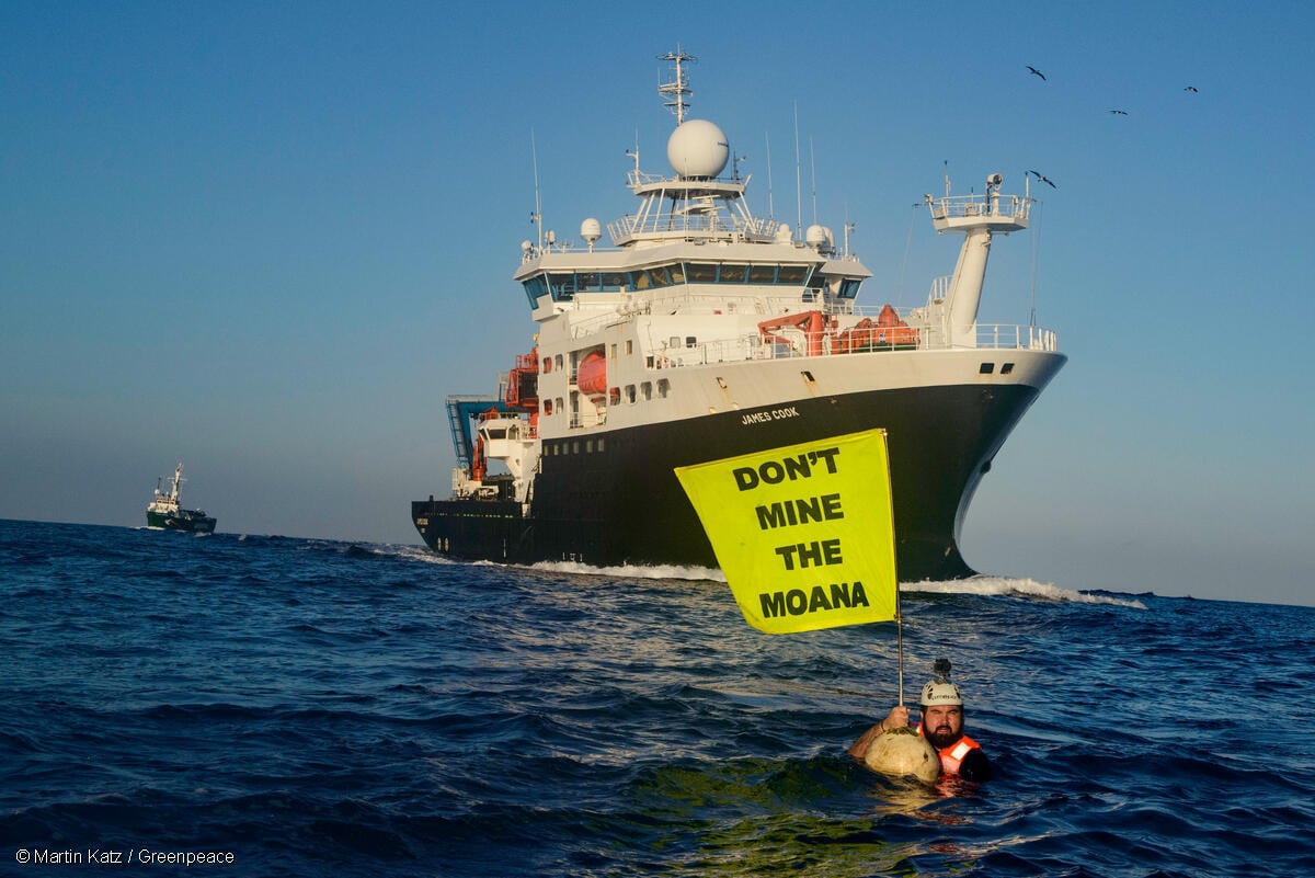 Activista de Greenpeace en medio del Oceáno Pacífico, sostiene una bandera en la que se lee: "No minen el Moana", frente al Buque Real de Investigación del Reino Unido.