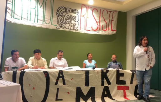 Personas sentadas en una mesa durante una conferencia de prensa, con una manta al frente que dice "No al tren mata porque el gobierno federal continúa violando y desacatando lo establecido en el Convenio 169 de la OIT.