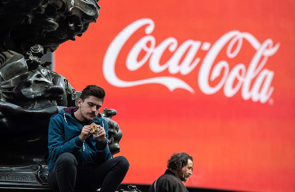 Fotografía de un hombre sentado frente a un banner gigante de Coca-Cola. Fotografía de un hombre sentado frente a un banner gigante de Coca-Cola.