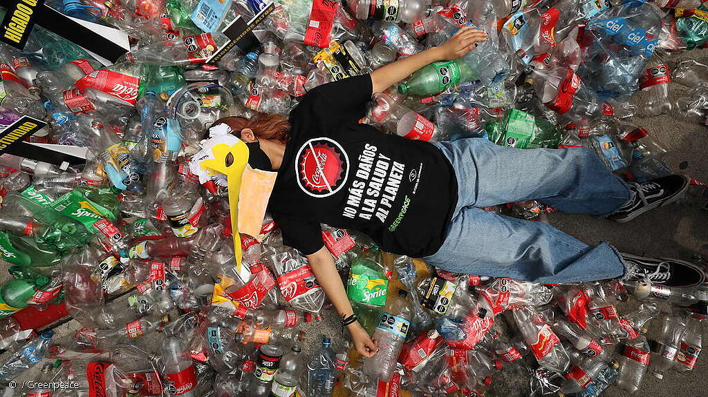 Activista de Greenpeace México recostada en el suelo sobre residuos plásticos de Coca-Cola durante manifestación pacífica.