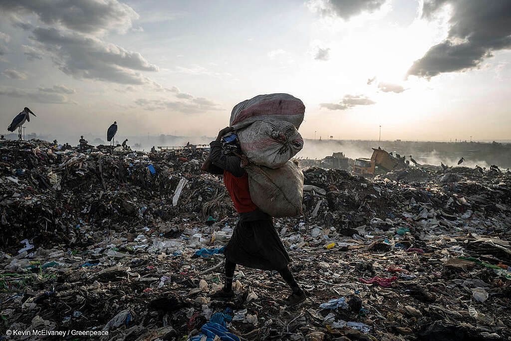Fotografía de un reciclador cargando un costal de residuos, mientras camina entre desiduos textiles y plásticos en el vertedero de Dandora, Nairobi.