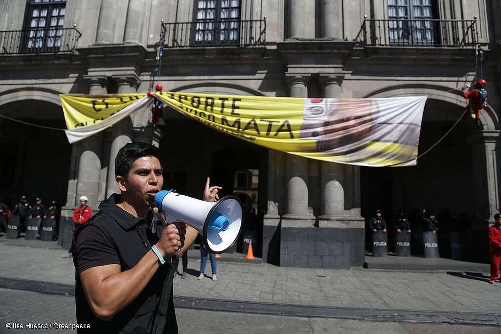 Retrato de Carlos Samayoa, campañista de Greenpeace México, sosteniendo un megáfono, detrás se observan activistas colgando una manta gigante en la que se lee "El transporte en el Estado de México mata".