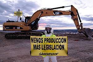 Activistas de Greenpeace en medio de un basurero, porta traje protección y sostiene una manta en la que se lee "Menos producción, más legislación".